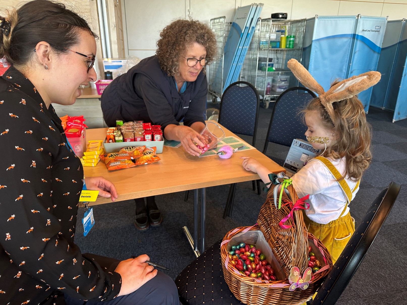 Etta at a blood donation session, standing by a snack table with two NHS Blood and Transplant staff members